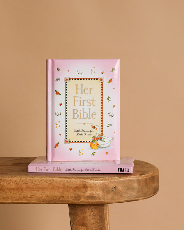Her First Bible," a pink children's book, stands upright on a wooden table with another copy underneath. The cover features animal and leaf illustrations, making it perfect for introducing God's word to a young girl's heart.