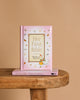 Her First Bible," a pink children's book, stands upright on a wooden table with another copy underneath. The cover features animal and leaf illustrations, making it perfect for introducing God's word to a young girl's heart.