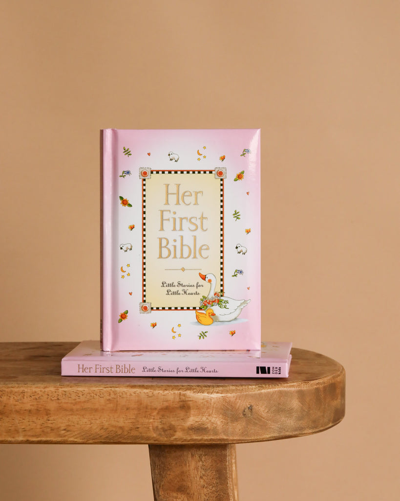 Her First Bible," a pink children's book, stands upright on a wooden table with another copy underneath. The cover features animal and leaf illustrations, making it perfect for introducing God's word to a young girl's heart.