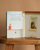 The "Just In Case You Ever Wonder Board Book" by Max Lucado rests open on a wooden stool, showing an illustration of a bear by a window and a poem about God's love, framed by a rooftop and soft blue sky.