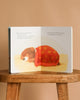 An open "Just In Case You Ever Wonder Board Book" rests on a wooden stool, displaying a brown bear cub sleeping under a red blanket, with comforting text about God's love on a soft, light background.