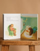 An open "Just In Case You Ever Wonder Board Book" rests on a wooden stool, displaying illustrations of a brown bear with a baby bear on the left and a larger bear watching over the crib on the right. Text appears beside the gentle artwork.
