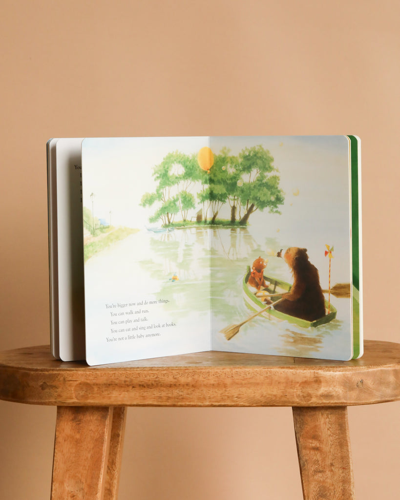 An open "Just In Case You Ever Wonder Board Book" by Max Lucado rests on a wooden stool, displaying an illustration of a bear and cub paddling a canoe beneath green trees and a bright sun, highlighting God's love in every scene.