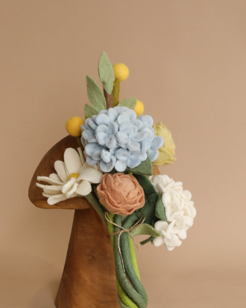 A decorative Flower Bouquet - Spring Bliss of eco-friendly felt flowers in soft pastel colors, including blue, white, peach, and green, mounted on a wooden stand with a beige background.