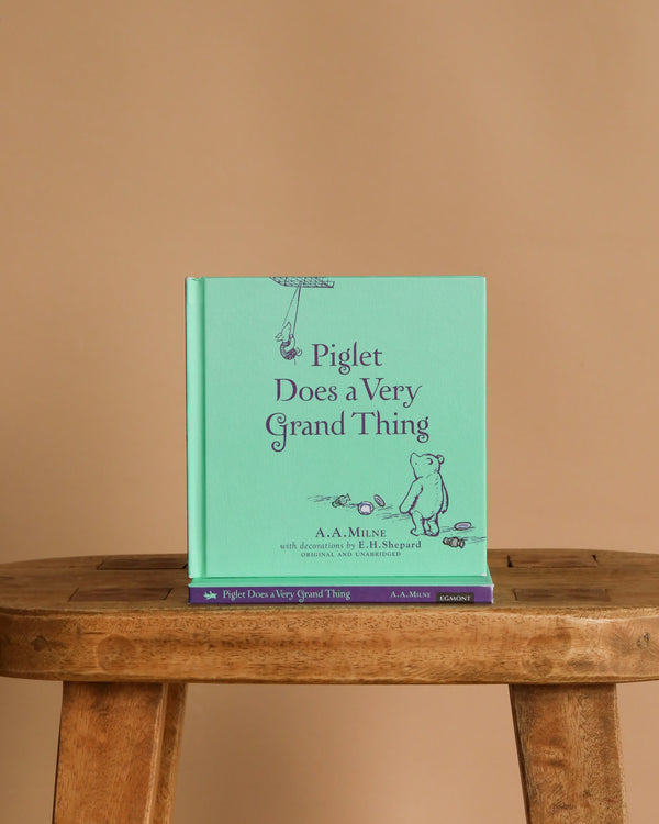 A mint green hardcover of "Winnie-the-Pooh: Piglet Does a Very Grand Thing" is displayed upright on a wooden stool against a plain beige background.