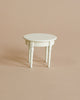 A small, round, white table with three legs sits on a beige background, ideal for showcasing your Maileg Pastry Chef Set With Grapat Wooden Bake Set.