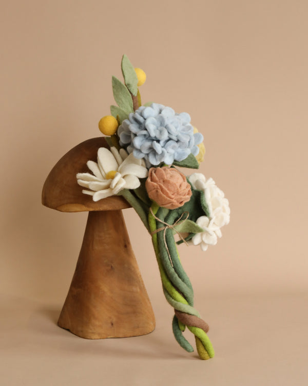 A wooden mushroom sculpture adorned with a Flower Bouquet - Spring Bliss in shades of blue, white, yellow, and peach, with green leaves and stems, set against a beige background.