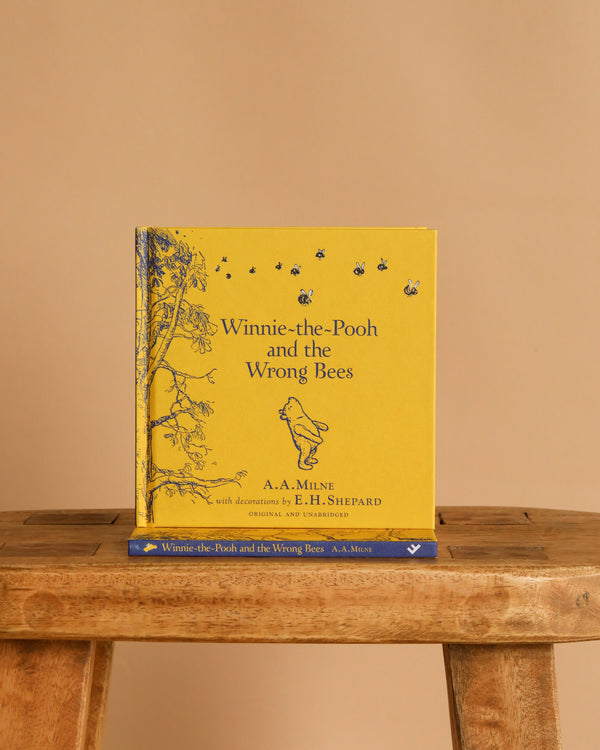 A yellow hardcover of "Winnie-the-Pooh: Winnie-the-Pooh and the Wrong Bees" stands upright on a wooden bench, featuring classic children’s story illustrations of Pooh, bees, and a tree, with another copy placed below it.
