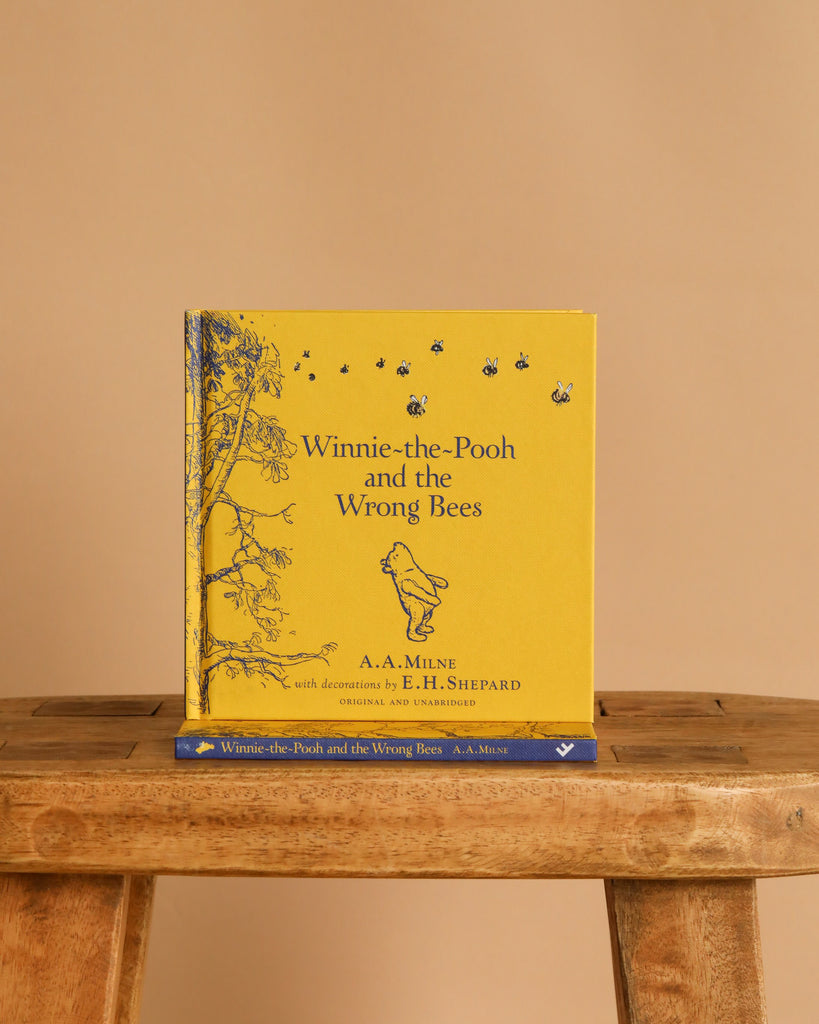 A yellow hardcover of "Winnie-the-Pooh: Winnie-the-Pooh and the Wrong Bees" stands upright on a wooden bench, featuring classic children’s story illustrations of Pooh, bees, and a tree, with another copy placed below it.
