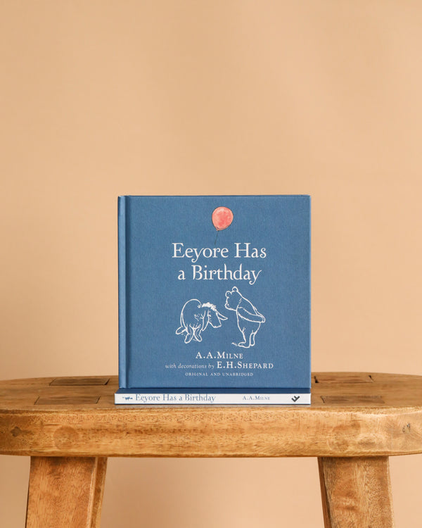 A blue hardcover titled "Winnie-the-Pooh: Eeyore Has A Birthday" stands upright on a wooden stool against a plain beige background, featuring simple line drawings of Eeyore and Winnie-the-Pooh from classic children's stories.