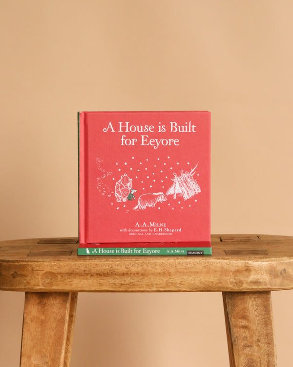 The red children's picture book "Winnie-the-Pooh: A House is Built for Eeyore" stands upright on a wooden stool, its cover showing a simple drawing of Winnie the Pooh and Eeyore against a beige background.