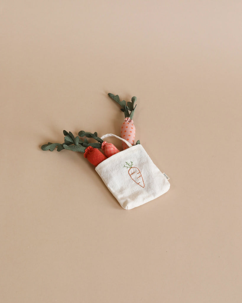 The Maileg Little Rabbit’s Carrot Harvest Set features a small white fabric bag embroidered with a carrot, containing fabric carrots and greenery, perfect for a Micro Rabbit or Maileg Extra Clothing.