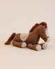 A Maileg Pony (ships in approximately one week) is shown lying down. It has a brown body with a textured saddle on its back that features a white and orange pattern. The mane and tail are made of brown yarn, and its muzzle and hooves are light gray. The background is a plain light beige, inviting the adventure of a lifetime.