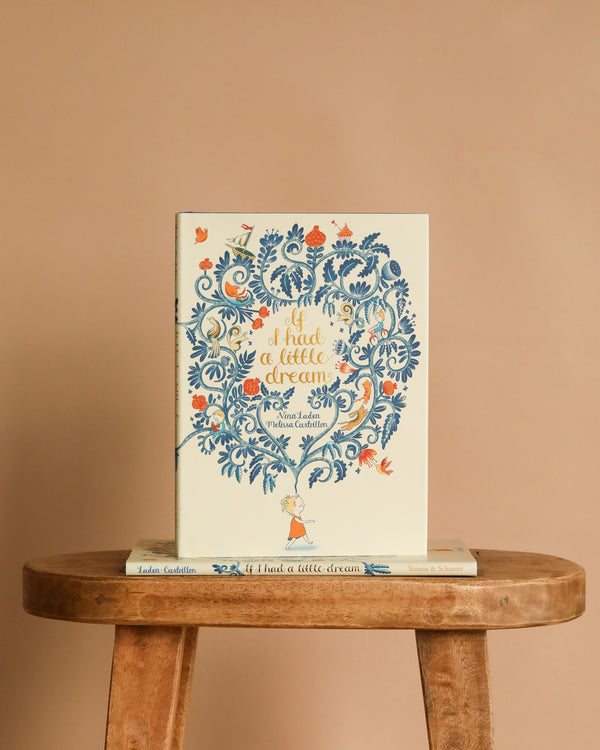 The picture book "If I Had a Little Dream by Nina Laden" with a floral cover and a small girl stands upright on a wooden stool, inviting readers to enjoy childhood's wonders, while another copy lies flat beneath it.