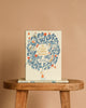 The picture book "If I Had a Little Dream by Nina Laden" with a floral cover and a small girl stands upright on a wooden stool, inviting readers to enjoy childhood's wonders, while another copy lies flat beneath it.