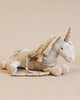 A Maileg Unicorn with a golden horn and multicolored yarn mane, made from recycled polyester, lies on a soft blanket against a light beige background.