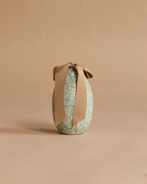 The Maileg Easter Egg, Small - Dusty Blue, featuring white floral designs and a large beige ribbon, stands upright on a plain beige background.