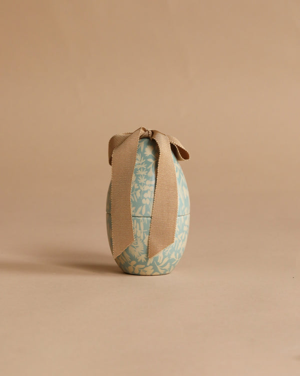 The Maileg Easter Egg, Small - Dusty Blue, featuring white floral designs and a large beige ribbon, stands upright on a plain beige background.