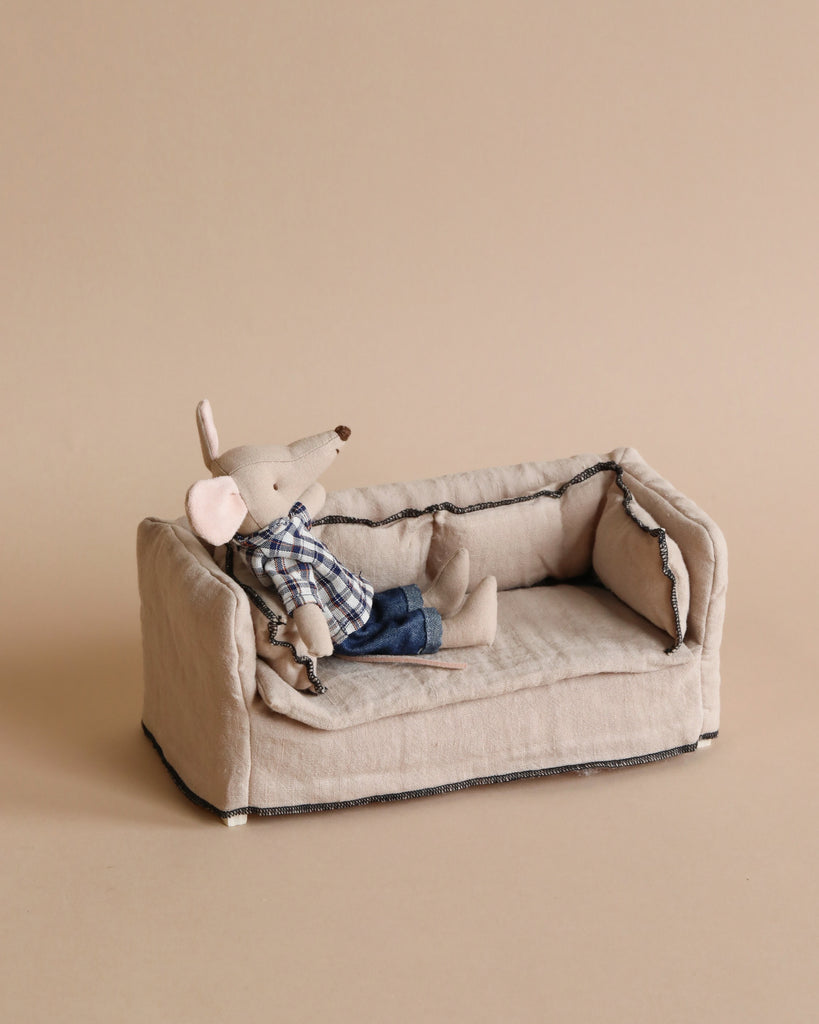 A Maileg dollhouse toy mouse wearing jeans and a plaid shirt lounges on a miniature Maileg couch, set against a plain beige background. The toy exudes a relaxed posture, sitting back