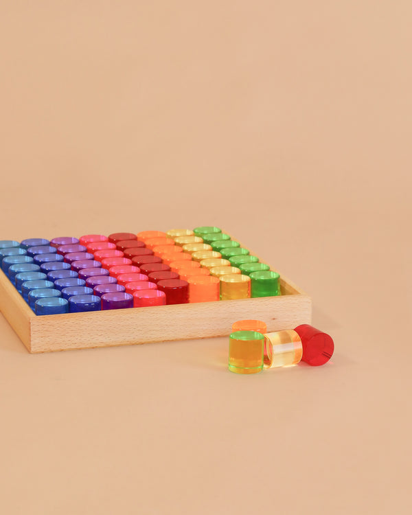 A Bauspiel Lucent Cylinders tray filled with vibrant, translucent cylinders in a rainbow pattern, with a yellow and a red cylinder placed outside the tray on a beige surface.