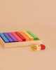 A Bauspiel Lucent Cylinders tray filled with vibrant, translucent cylinders in a rainbow pattern, with a yellow and a red cylinder placed outside the tray on a beige surface.