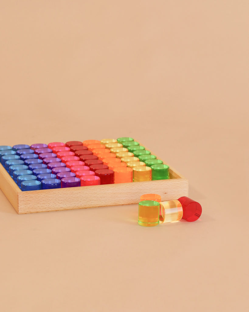 A Bauspiel Lucent Cylinders tray filled with vibrant, translucent cylinders in a rainbow pattern, with a yellow and a red cylinder placed outside the tray on a beige surface.