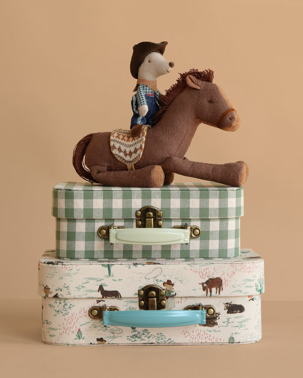 A Maileg Cowboy Set features a cowboy mouse on a plush rocking horse, displayed atop two Maileg suitcases—one green plaid, one with a pastoral print—arranged against a beige background.