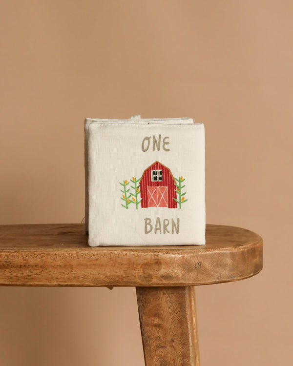 The Little Farm Soft Book for infants features "ONE BARN" and a red barn on the cover, displayed on a wooden bench with a beige background—a charming farm-themed gift.