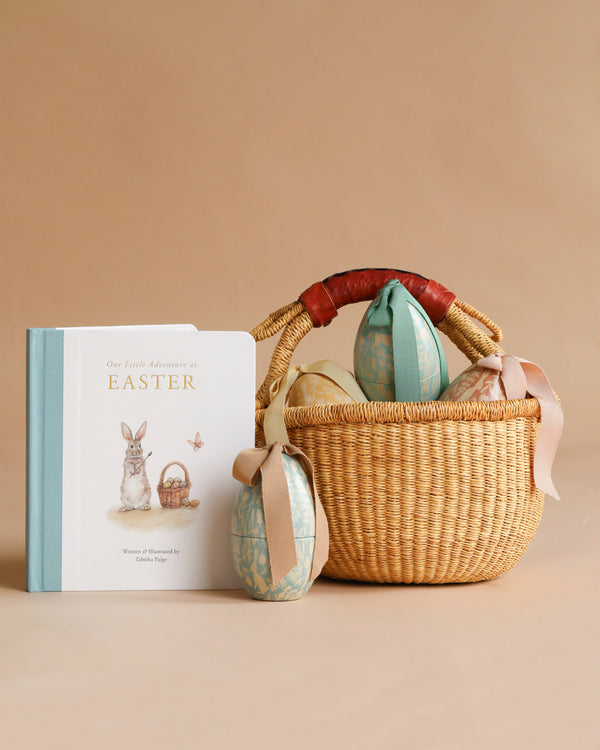 The Maileg Easter Basket Set features a woven handmade basket with a brown handle filled with decorated eggs and includes a bunny-themed board book for toddlers. Extra ribbon-tied eggs are displayed beside the basket on a beige background.