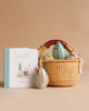 The Maileg Easter Basket Set features a woven handmade basket with a brown handle filled with decorated eggs and includes a bunny-themed board book for toddlers. Extra ribbon-tied eggs are displayed beside the basket on a beige background.