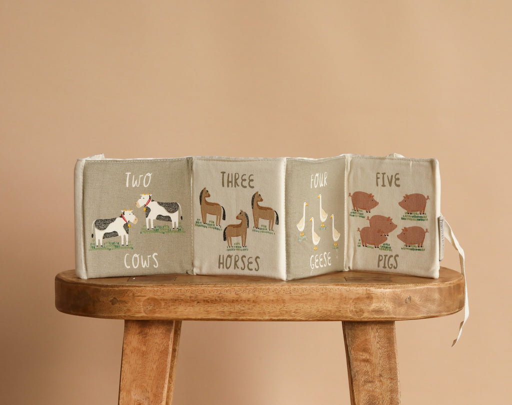 The Little Farm Soft Book displays charming illustrations of two cows, three horses, four geese, and five pigs on a wooden stool with a beige backdrop—ideal as an infant book or thoughtful baby gift.