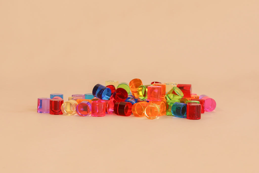 A pile of Bauspiel Lucent Cylinders—colorful, translucent plastic pieces—scattered on a beige surface with a plain beige background, evoking the vibrant, creative look of popular construction sets.