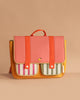 The Sticky Lemon School Bag in Coral Pink and Submarine Yellow, made of waterproof nylon, features a colorful design with an orange body, pink flap, red straps, and striped front pockets against a beige background.