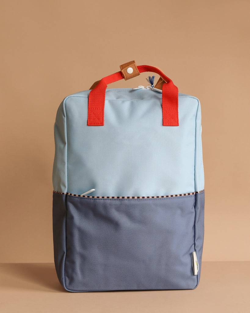 A Sticky Lemon Backpack Large in Bissou Blue + Blue Whale stands upright against a beige background, featuring a color-blocked design, red handles, front pocket, and a small brown tab with white button. Made from recycled PET.