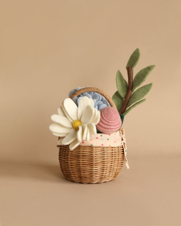 A small wicker basket filled with eco-friendly, handmade flowers and plants, including a white daisy, pink rose, light blue knit flower, and green leaves. The arrangement sits against a beige background. This is the Basket of Felt Flowers.