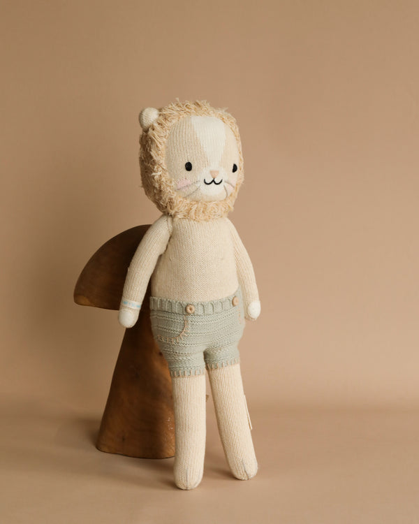A Cuddle + Kind Sawyer The Lion - Large with a smiling face, wearing a knitted cream sweater made from Peruvian cotton yarn and green shorts, stands against a soft beige background.
