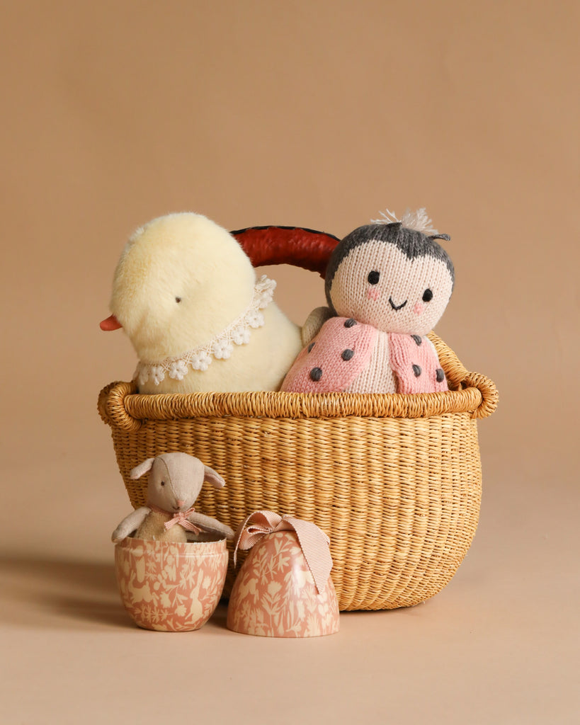 The Maileg Easter Basket Set features a woven basket holding a plush yellow duck and a knitted doll, with a Maileg Easter Egg beside them. In front, a fabric bunny and two pink patterned eggs rest on the beige background.