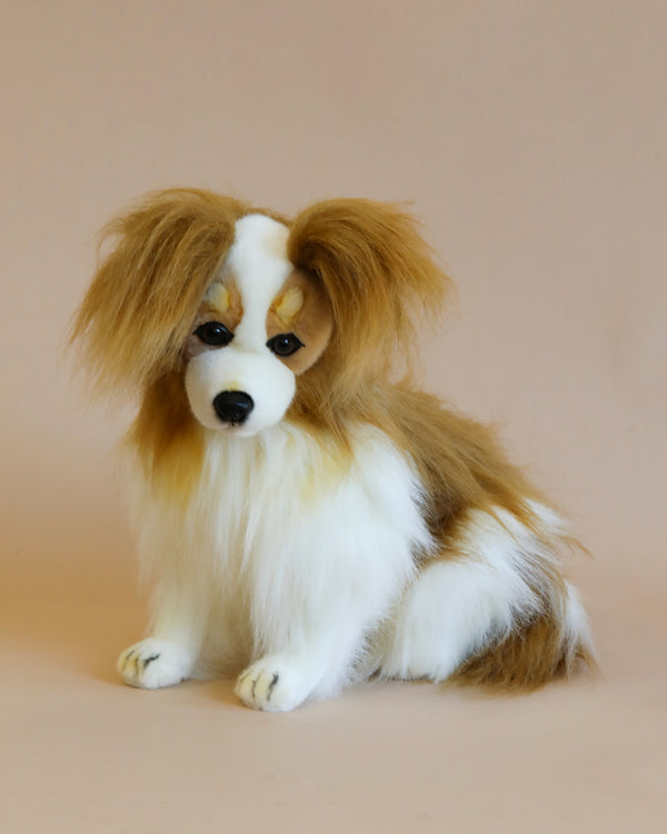 A lifelike Papillon Dog Stuffed Animal with fluffy tan and white fur, big floppy ears, and expressive brown eyes, handcrafted by artisans and sitting against a pale pink background.