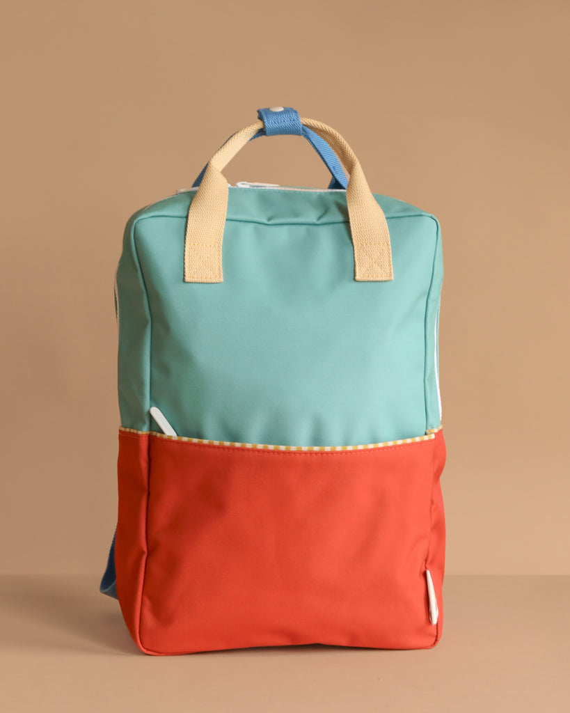 The Sticky Lemon Backpack Large | Color Blocking | Aquatic Green + Shipwreck Rust features a teal upper, red lower section, beige handles, blue strap accents, and is made from recycled PET with a waterproof lining. Shown upright on a plain beige background.