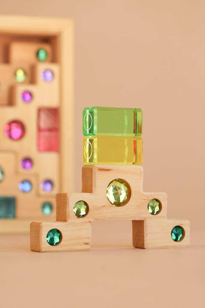 The Bauspiel Colored Staircase with Luminous Blocks - 32 Piece is stacked in a small arch shape in front, with more blocks arranged in a wooden frame behind, creating an inviting and colorful construction set against a beige backdrop.
