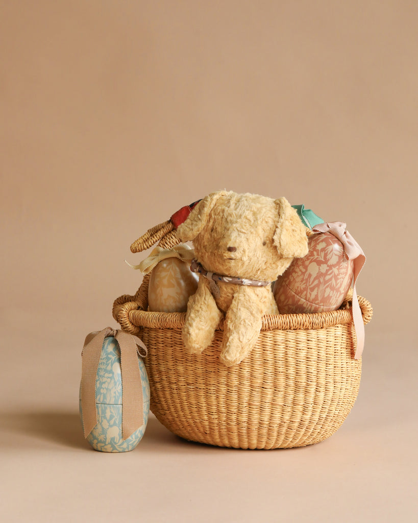 A Maileg Easter Basket Set sits against a plain beige background, featuring a plush Maileg Puppy and several patterned fabric-wrapped items. Beside it rests another fabric-wrapped Maileg Easter Egg.