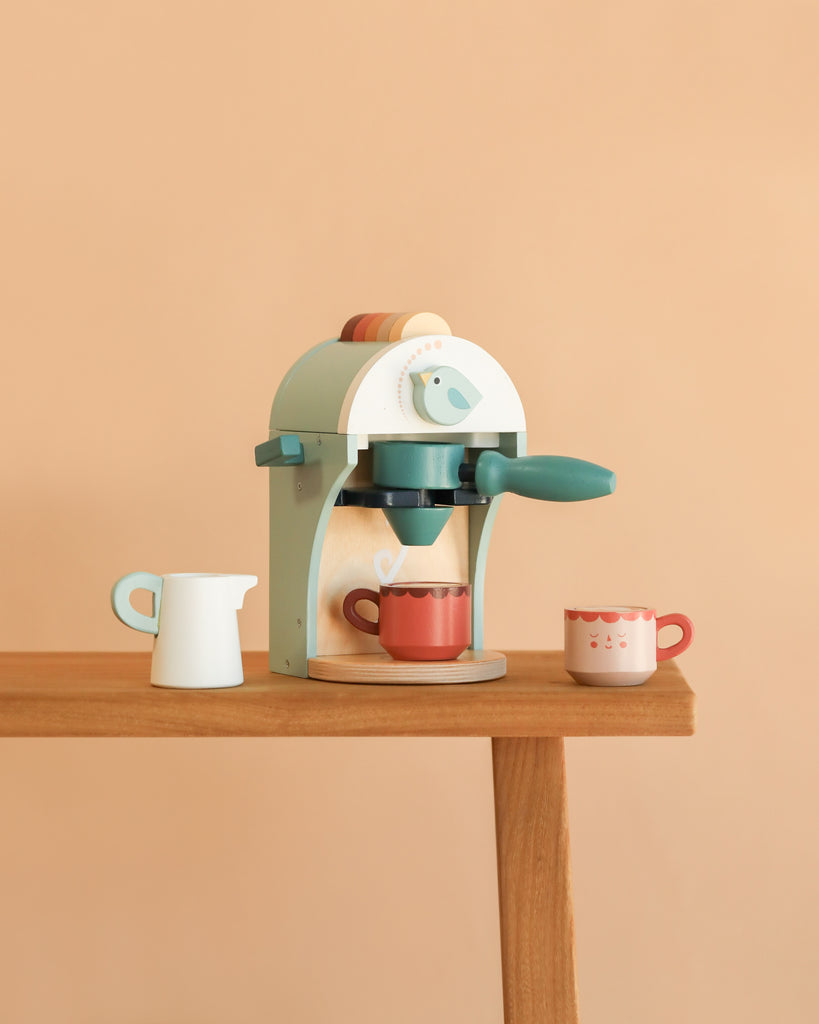 The Babyccino Maker, with its blue handle, sits on a table alongside a white toy milk jug and a red cup, bringing the charm of Bird’s Nest Café to your child’s imaginative play against a peach-colored background.