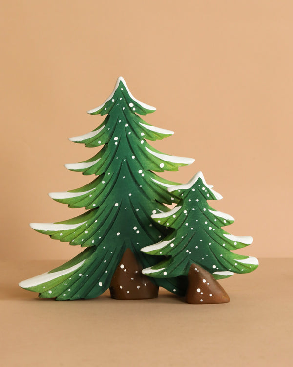 The Bumbu Handmade Wooden Snowy Fir Tree Set features two green, snow-dusted fir trees crafted from sustainably sourced wood, standing side by side on a beige surface with brown stones—a delightful Montessori toy.