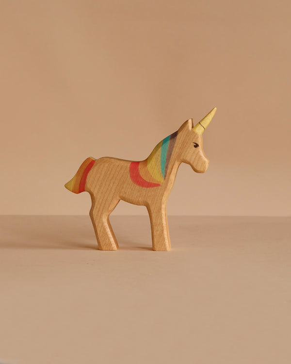 The Handmade Holzwald Unicorn Rainbow, a sustainable wooden toy with a yellow horn and rainbow mane, stands on a beige surface against a matching beige background.