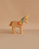 The Handmade Holzwald Unicorn Rainbow, a sustainable wooden toy with a yellow horn and rainbow mane, stands on a beige surface against a matching beige background.