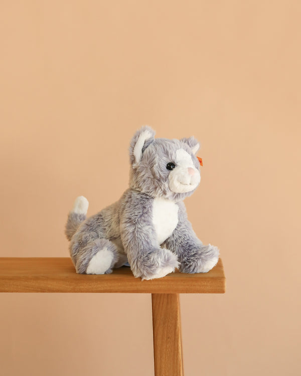 A Steiff Cassie Cat plush in ice blue sits on the edge of a wooden bench against a plain beige background.