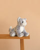 A Steiff Cassie Cat plush in ice blue sits on the edge of a wooden bench against a plain beige background.