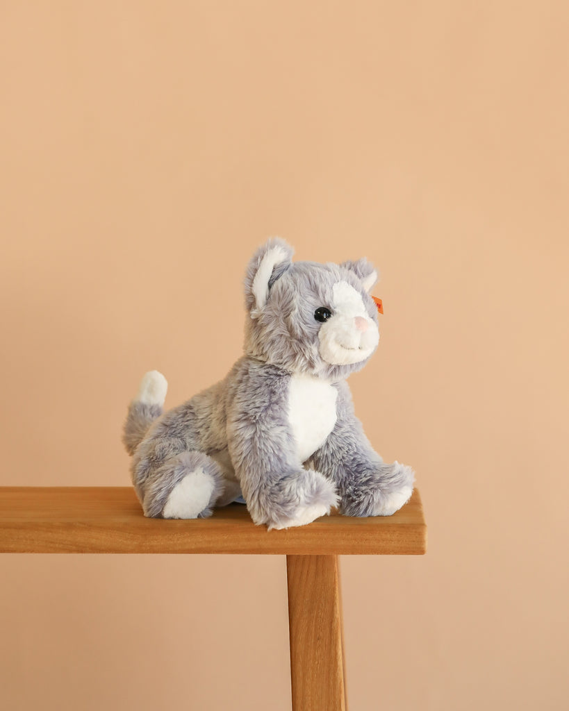 A Steiff Cassie Cat plush in ice blue sits on the edge of a wooden bench against a plain beige background.