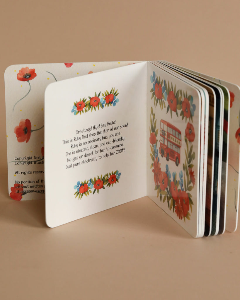 An open children's board book with illustrated red flowers on its pages, positioned on a beige background. The visible page contains a short story about Ruby The Red Electric Bus Book.