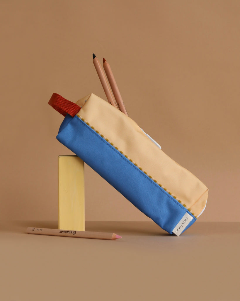 The Sticky Lemon Pencil Case | Color Blocking | Sandy Yellow + Radar Blue, made from recycled PET bottles, stands upright on a yellow block with two pencils inside and one colored pencil lying on a beige surface. The background is solid light brown.